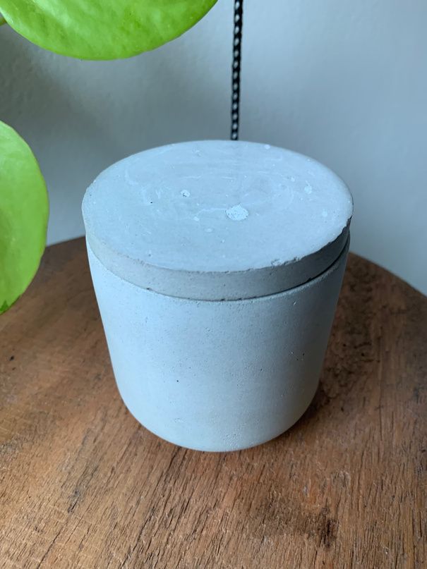 Vela Marie: pote de concreto cinza com tampa, sobre uma mesa de madeira
